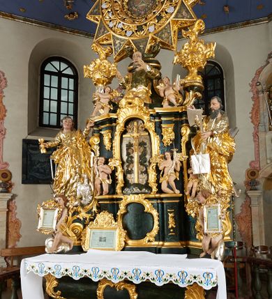 Zdjęcie nr 12: Ołtarz dwustronny, sarkofagowy, dekorowany rocaille'm, mensa drewniana na planie kwadratu, od frontu z kamiennym portatylem.
Nastawa ołtarzowa dwustronna, na rzucie leżącej litery X, w formie  rozbudowanego tabernakulum z tronem wystawienia. Obie strony są analogiczne pod względem architektonicznym. Tabernakulum na cokole, o wypukłej ściance frontowej, flankowanej parą pilastrów o trzonach dekorowanym płycinami z rocaille'm, dźwigających gzyms wieńczący. W polu drzwiczki ujęte rokokowym kartuszem. Na tabernakulum tron wystawienia o wklęsło-wypukłym licu, ujęty parą impostów podtrzymywanych przez rocaille'owe konsole oraz pilastrami w narożach, których trzony przyjmują formę spływów wolutowych, sięgające podstawy tabernakulum, na których stoją figury. W polu środkowym tronu wystawienia znajduje się obrotowa nisza o nieregularnym kształcie ujęta obramieniem z rocaille'u, przesłonięta wypukłą ścianką z płaskorzeźbionym krucyfiksem  i winnym gronem, zwieńczona rzeźbioną półpostacią. Całość wieńczy cokół z dwoma ramionami, na których stoją rocaille'owe wazony. Na cokole osadzona jest dużych rozmiarów, dwustronna, dwunastoramienna gwiazda relikwiarzowa, miażdżąca węża. Gwiazda z obu stron ujęta jest kilkoma pierścieniami z winnego grona oraz ornamentu roślinnego, w ramionach oraz w polu głównym umieszczone są za szkłem relikwie wśród kwiatowych dekoracji. 
Na awersie ołtarza, na mensie znajdują się dwie figurki puttów siedzących na obłokach i trzymających tabliczki z kanonami. Całość flankują ustawione na spływach wolutowych figury św. Jana Ewangelisty i św. Bartłomieja, tron wieńczy półpostać Boga Ojca, ujęta przez dwa putta z otwartymi księgami. Obrotową niszę flankują dwa aniołki trzymające lichtarze. Na rewersie ołtarza na spływach wolutowych stoją figury św. Andrzeja i św. Piotra, tron wieńczy półpostać Mojżesza flankowana przez dwa putta z otwartymi księgami.
Struktura polichromowana w kolorze ciemnozielonym, ornamentyka i profile złocone, detal srebrzony.
