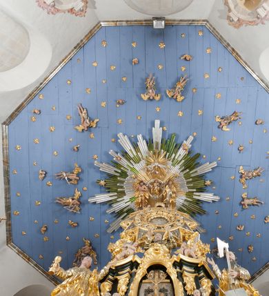 Zdjęcie nr 18: Drewniany, polichromowany na niebiesko strop, na planie ośmioboku, ujęty profilowaną ramą. W jego centrum grupa Koronacji Marii w glorii promienistej. W środku grupy figura Matki Boskiej na tle opaski dekorowanej kwiatami. Maria ukazana frontalnie, w kontrapoście, z rękami złożonymi w geście modlitwy. Twarz ma pociągłą, szczupłą, o młodzieńczych rysach, okoloną długimi włosami, upiętymi z tyłu głowy. Ubrana jest w srebrzoną suknię oraz złocony płaszcz narzucony na ramiona i zawinięty na lewej nodze, na głowie ma koronę zamkniętą. Marię flankują dwie banderole z napisem: „FIAT”. Po jej prawej znacznie większa figura Chrystusa, ukazanego frontalnie, w pozycji siedzącej, z głową skierowaną do wewnątrz; jego lewa ręka spoczywa na piersi, prawa jest ugięta i uniesiona w geście wskazującym. Twarz ma pociągłą, szczupłą, okoloną brodą skręconą w dwa pukle, oraz kręconymi włosami. Chrystus przewiązany jest w partii bioder złoconym płaszczem otaczającym postać i odsłaniającym lewą nogę. Jego ciało w odsłoniętych partiach, ma delikatnie zarysowaną muskulaturę, z zaznaczonymi śladami męki. Po lewej stronie Marii znacznie większa figura Boga Ojca, ukazanego frontalnie, w pozycji siedzącej. Lewą ręką przytrzymuje jabłko królewskie, w dłoni dzierży berło, prawą wyciąga w geście błogosławieństwa. Twarz ma pociągłą, o starczych rysach, okoloną długą, kręconą brodą opadającą na pierś oraz krótkimi włosami. Ubrany jest w obficie drapowaną srebrzoną tunikę, przepasaną w talii oraz złocony płaszcz z dekoracyjną, kameryzowaną lamówką, zapięty na piersi i opadający na boki, z zawiniętym fragmentem tkaniny na lewej nodze. Nad postaciami nadlatująca gołębica Ducha Świętego w glorii promienistej z banderolą z napisem: „FIAT”. Grupa na tle trzystrefowej glorii promienistej dekorowanej gwiazdami i uskrzydlonymi główkami anielskimi. Glorię z grupą Koronacji otacza dwunastu aniołów, ułożonych naprzemiennie w cztery pary muzykujących postaci (ze skrzypcami, wiolonczelą, gitarą, trąbką, harfą) z czterema aniołami podtrzymującymi tace z symbolicznymi przedmiotami (różaniec, wieniec laurowy, chleb (?), dyscyplina). Strop dekorowany jest w całości gwiazdami i uskrzydlonymi główkami anielskimi. Figury w partiach ciała polichromowane są naturalistycznie, ich szaty są złocone, srebrzone i laserowane; atrybuty są srebrzone. 

