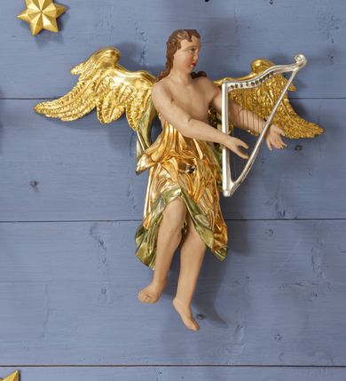 Zdjęcie nr 9: Drewniany, polichromowany na niebiesko strop, na planie ośmioboku, ujęty profilowaną ramą. W jego centrum grupa Koronacji Marii w glorii promienistej. W środku grupy figura Matki Boskiej na tle opaski dekorowanej kwiatami. Maria ukazana frontalnie, w kontrapoście, z rękami złożonymi w geście modlitwy. Twarz ma pociągłą, szczupłą, o młodzieńczych rysach, okoloną długimi włosami, upiętymi z tyłu głowy. Ubrana jest w srebrzoną suknię oraz złocony płaszcz narzucony na ramiona i zawinięty na lewej nodze, na głowie ma koronę zamkniętą. Marię flankują dwie banderole z napisem: „FIAT”. Po jej prawej znacznie większa figura Chrystusa, ukazanego frontalnie, w pozycji siedzącej, z głową skierowaną do wewnątrz; jego lewa ręka spoczywa na piersi, prawa jest ugięta i uniesiona w geście wskazującym. Twarz ma pociągłą, szczupłą, okoloną brodą skręconą w dwa pukle, oraz kręconymi włosami. Chrystus przewiązany jest w partii bioder złoconym płaszczem otaczającym postać i odsłaniającym lewą nogę. Jego ciało w odsłoniętych partiach, ma delikatnie zarysowaną muskulaturę, z zaznaczonymi śladami męki. Po lewej stronie Marii znacznie większa figura Boga Ojca, ukazanego frontalnie, w pozycji siedzącej. Lewą ręką przytrzymuje jabłko królewskie, w dłoni dzierży berło, prawą wyciąga w geście błogosławieństwa. Twarz ma pociągłą, o starczych rysach, okoloną długą, kręconą brodą opadającą na pierś oraz krótkimi włosami. Ubrany jest w obficie drapowaną srebrzoną tunikę, przepasaną w talii oraz złocony płaszcz z dekoracyjną, kameryzowaną lamówką, zapięty na piersi i opadający na boki, z zawiniętym fragmentem tkaniny na lewej nodze. Nad postaciami nadlatująca gołębica Ducha Świętego w glorii promienistej z banderolą z napisem: „FIAT”. Grupa na tle trzystrefowej glorii promienistej dekorowanej gwiazdami i uskrzydlonymi główkami anielskimi. Glorię z grupą Koronacji otacza dwunastu aniołów, ułożonych naprzemiennie w cztery pary muzykujących postaci (ze skrzypcami, wiolonczelą, gitarą, trąbką, harfą) z czterema aniołami podtrzymującymi tace z symbolicznymi przedmiotami (różaniec, wieniec laurowy, chleb (?), dyscyplina). Strop dekorowany jest w całości gwiazdami i uskrzydlonymi główkami anielskimi. Figury w partiach ciała polichromowane są naturalistycznie, ich szaty są złocone, srebrzone i laserowane; atrybuty są srebrzone. 

