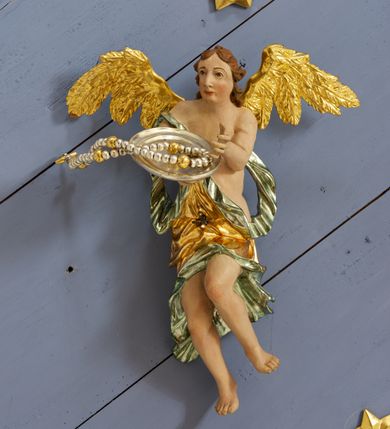 Zdjęcie nr 14: Drewniany, polichromowany na niebiesko strop, na planie ośmioboku, ujęty profilowaną ramą. W jego centrum grupa Koronacji Marii w glorii promienistej. W środku grupy figura Matki Boskiej na tle opaski dekorowanej kwiatami. Maria ukazana frontalnie, w kontrapoście, z rękami złożonymi w geście modlitwy. Twarz ma pociągłą, szczupłą, o młodzieńczych rysach, okoloną długimi włosami, upiętymi z tyłu głowy. Ubrana jest w srebrzoną suknię oraz złocony płaszcz narzucony na ramiona i zawinięty na lewej nodze, na głowie ma koronę zamkniętą. Marię flankują dwie banderole z napisem: „FIAT”. Po jej prawej znacznie większa figura Chrystusa, ukazanego frontalnie, w pozycji siedzącej, z głową skierowaną do wewnątrz; jego lewa ręka spoczywa na piersi, prawa jest ugięta i uniesiona w geście wskazującym. Twarz ma pociągłą, szczupłą, okoloną brodą skręconą w dwa pukle, oraz kręconymi włosami. Chrystus przewiązany jest w partii bioder złoconym płaszczem otaczającym postać i odsłaniającym lewą nogę. Jego ciało w odsłoniętych partiach, ma delikatnie zarysowaną muskulaturę, z zaznaczonymi śladami męki. Po lewej stronie Marii znacznie większa figura Boga Ojca, ukazanego frontalnie, w pozycji siedzącej. Lewą ręką przytrzymuje jabłko królewskie, w dłoni dzierży berło, prawą wyciąga w geście błogosławieństwa. Twarz ma pociągłą, o starczych rysach, okoloną długą, kręconą brodą opadającą na pierś oraz krótkimi włosami. Ubrany jest w obficie drapowaną srebrzoną tunikę, przepasaną w talii oraz złocony płaszcz z dekoracyjną, kameryzowaną lamówką, zapięty na piersi i opadający na boki, z zawiniętym fragmentem tkaniny na lewej nodze. Nad postaciami nadlatująca gołębica Ducha Świętego w glorii promienistej z banderolą z napisem: „FIAT”. Grupa na tle trzystrefowej glorii promienistej dekorowanej gwiazdami i uskrzydlonymi główkami anielskimi. Glorię z grupą Koronacji otacza dwunastu aniołów, ułożonych naprzemiennie w cztery pary muzykujących postaci (ze skrzypcami, wiolonczelą, gitarą, trąbką, harfą) z czterema aniołami podtrzymującymi tace z symbolicznymi przedmiotami (różaniec, wieniec laurowy, chleb (?), dyscyplina). Strop dekorowany jest w całości gwiazdami i uskrzydlonymi główkami anielskimi. Figury w partiach ciała polichromowane są naturalistycznie, ich szaty są złocone, srebrzone i laserowane; atrybuty są srebrzone. 

