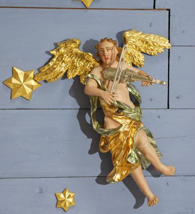 Zdjęcie nr 15: Drewniany, polichromowany na niebiesko strop, na planie ośmioboku, ujęty profilowaną ramą. W jego centrum grupa Koronacji Marii w glorii promienistej. W środku grupy figura Matki Boskiej na tle opaski dekorowanej kwiatami. Maria ukazana frontalnie, w kontrapoście, z rękami złożonymi w geście modlitwy. Twarz ma pociągłą, szczupłą, o młodzieńczych rysach, okoloną długimi włosami, upiętymi z tyłu głowy. Ubrana jest w srebrzoną suknię oraz złocony płaszcz narzucony na ramiona i zawinięty na lewej nodze, na głowie ma koronę zamkniętą. Marię flankują dwie banderole z napisem: „FIAT”. Po jej prawej znacznie większa figura Chrystusa, ukazanego frontalnie, w pozycji siedzącej, z głową skierowaną do wewnątrz; jego lewa ręka spoczywa na piersi, prawa jest ugięta i uniesiona w geście wskazującym. Twarz ma pociągłą, szczupłą, okoloną brodą skręconą w dwa pukle, oraz kręconymi włosami. Chrystus przewiązany jest w partii bioder złoconym płaszczem otaczającym postać i odsłaniającym lewą nogę. Jego ciało w odsłoniętych partiach, ma delikatnie zarysowaną muskulaturę, z zaznaczonymi śladami męki. Po lewej stronie Marii znacznie większa figura Boga Ojca, ukazanego frontalnie, w pozycji siedzącej. Lewą ręką przytrzymuje jabłko królewskie, w dłoni dzierży berło, prawą wyciąga w geście błogosławieństwa. Twarz ma pociągłą, o starczych rysach, okoloną długą, kręconą brodą opadającą na pierś oraz krótkimi włosami. Ubrany jest w obficie drapowaną srebrzoną tunikę, przepasaną w talii oraz złocony płaszcz z dekoracyjną, kameryzowaną lamówką, zapięty na piersi i opadający na boki, z zawiniętym fragmentem tkaniny na lewej nodze. Nad postaciami nadlatująca gołębica Ducha Świętego w glorii promienistej z banderolą z napisem: „FIAT”. Grupa na tle trzystrefowej glorii promienistej dekorowanej gwiazdami i uskrzydlonymi główkami anielskimi. Glorię z grupą Koronacji otacza dwunastu aniołów, ułożonych naprzemiennie w cztery pary muzykujących postaci (ze skrzypcami, wiolonczelą, gitarą, trąbką, harfą) z czterema aniołami podtrzymującymi tace z symbolicznymi przedmiotami (różaniec, wieniec laurowy, chleb (?), dyscyplina). Strop dekorowany jest w całości gwiazdami i uskrzydlonymi główkami anielskimi. Figury w partiach ciała polichromowane są naturalistycznie, ich szaty są złocone, srebrzone i laserowane; atrybuty są srebrzone. 

