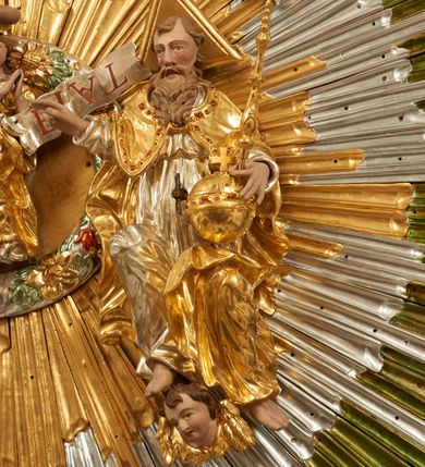 Zdjęcie nr 17: Drewniany, polichromowany na niebiesko strop, na planie ośmioboku, ujęty profilowaną ramą. W jego centrum grupa Koronacji Marii w glorii promienistej. W środku grupy figura Matki Boskiej na tle opaski dekorowanej kwiatami. Maria ukazana frontalnie, w kontrapoście, z rękami złożonymi w geście modlitwy. Twarz ma pociągłą, szczupłą, o młodzieńczych rysach, okoloną długimi włosami, upiętymi z tyłu głowy. Ubrana jest w srebrzoną suknię oraz złocony płaszcz narzucony na ramiona i zawinięty na lewej nodze, na głowie ma koronę zamkniętą. Marię flankują dwie banderole z napisem: „FIAT”. Po jej prawej znacznie większa figura Chrystusa, ukazanego frontalnie, w pozycji siedzącej, z głową skierowaną do wewnątrz; jego lewa ręka spoczywa na piersi, prawa jest ugięta i uniesiona w geście wskazującym. Twarz ma pociągłą, szczupłą, okoloną brodą skręconą w dwa pukle, oraz kręconymi włosami. Chrystus przewiązany jest w partii bioder złoconym płaszczem otaczającym postać i odsłaniającym lewą nogę. Jego ciało w odsłoniętych partiach, ma delikatnie zarysowaną muskulaturę, z zaznaczonymi śladami męki. Po lewej stronie Marii znacznie większa figura Boga Ojca, ukazanego frontalnie, w pozycji siedzącej. Lewą ręką przytrzymuje jabłko królewskie, w dłoni dzierży berło, prawą wyciąga w geście błogosławieństwa. Twarz ma pociągłą, o starczych rysach, okoloną długą, kręconą brodą opadającą na pierś oraz krótkimi włosami. Ubrany jest w obficie drapowaną srebrzoną tunikę, przepasaną w talii oraz złocony płaszcz z dekoracyjną, kameryzowaną lamówką, zapięty na piersi i opadający na boki, z zawiniętym fragmentem tkaniny na lewej nodze. Nad postaciami nadlatująca gołębica Ducha Świętego w glorii promienistej z banderolą z napisem: „FIAT”. Grupa na tle trzystrefowej glorii promienistej dekorowanej gwiazdami i uskrzydlonymi główkami anielskimi. Glorię z grupą Koronacji otacza dwunastu aniołów, ułożonych naprzemiennie w cztery pary muzykujących postaci (ze skrzypcami, wiolonczelą, gitarą, trąbką, harfą) z czterema aniołami podtrzymującymi tace z symbolicznymi przedmiotami (różaniec, wieniec laurowy, chleb (?), dyscyplina). Strop dekorowany jest w całości gwiazdami i uskrzydlonymi główkami anielskimi. Figury w partiach ciała polichromowane są naturalistycznie, ich szaty są złocone, srebrzone i laserowane; atrybuty są srebrzone. 

