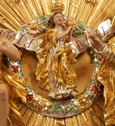 Zdjęcie nr 16: Drewniany, polichromowany na niebiesko strop, na planie ośmioboku, ujęty profilowaną ramą. W jego centrum grupa Koronacji Marii w glorii promienistej. W środku grupy figura Matki Boskiej na tle opaski dekorowanej kwiatami. Maria ukazana frontalnie, w kontrapoście, z rękami złożonymi w geście modlitwy. Twarz ma pociągłą, szczupłą, o młodzieńczych rysach, okoloną długimi włosami, upiętymi z tyłu głowy. Ubrana jest w srebrzoną suknię oraz złocony płaszcz narzucony na ramiona i zawinięty na lewej nodze, na głowie ma koronę zamkniętą. Marię flankują dwie banderole z napisem: „FIAT”. Po jej prawej znacznie większa figura Chrystusa, ukazanego frontalnie, w pozycji siedzącej, z głową skierowaną do wewnątrz; jego lewa ręka spoczywa na piersi, prawa jest ugięta i uniesiona w geście wskazującym. Twarz ma pociągłą, szczupłą, okoloną brodą skręconą w dwa pukle, oraz kręconymi włosami. Chrystus przewiązany jest w partii bioder złoconym płaszczem otaczającym postać i odsłaniającym lewą nogę. Jego ciało w odsłoniętych partiach, ma delikatnie zarysowaną muskulaturę, z zaznaczonymi śladami męki. Po lewej stronie Marii znacznie większa figura Boga Ojca, ukazanego frontalnie, w pozycji siedzącej. Lewą ręką przytrzymuje jabłko królewskie, w dłoni dzierży berło, prawą wyciąga w geście błogosławieństwa. Twarz ma pociągłą, o starczych rysach, okoloną długą, kręconą brodą opadającą na pierś oraz krótkimi włosami. Ubrany jest w obficie drapowaną srebrzoną tunikę, przepasaną w talii oraz złocony płaszcz z dekoracyjną, kameryzowaną lamówką, zapięty na piersi i opadający na boki, z zawiniętym fragmentem tkaniny na lewej nodze. Nad postaciami nadlatująca gołębica Ducha Świętego w glorii promienistej z banderolą z napisem: „FIAT”. Grupa na tle trzystrefowej glorii promienistej dekorowanej gwiazdami i uskrzydlonymi główkami anielskimi. Glorię z grupą Koronacji otacza dwunastu aniołów, ułożonych naprzemiennie w cztery pary muzykujących postaci (ze skrzypcami, wiolonczelą, gitarą, trąbką, harfą) z czterema aniołami podtrzymującymi tace z symbolicznymi przedmiotami (różaniec, wieniec laurowy, chleb (?), dyscyplina). Strop dekorowany jest w całości gwiazdami i uskrzydlonymi główkami anielskimi. Figury w partiach ciała polichromowane są naturalistycznie, ich szaty są złocone, srebrzone i laserowane; atrybuty są srebrzone. 

