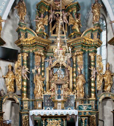 Zdjęcie nr 5: Nastawa ołtarzowa architektoniczna, trójosiowa z bramkami, dwukondygnacyjna, na dwustrefowym cokole. Ołtarz kamienny, prostopadłościenny, w drewnianej obudowie. Antepedium w formie wydłużonego, leżącego prostokąta, w polu obraz Ostatniej Wieczerzy. Mensa drewniana. Osie nastawy wydzielone parą półfilarów ujętych dwiema parami kolumn o kapitelach kompozytowych i skręconych trzonach. Dźwigają one pełne belkowanie silnie wyłamane na osi filarów, z mocno wysuniętym gzymsem, na skraju belkowania przerwany półkolisty przyczółek zwrócony na zewnątrz, na nim cokoły z figurami św. Władysława i św. Stefana. Na tle filarów ustawione figury św. Augustyna i św. Wojciecha, na bramkach rzeźby św. Marcina i św. Jana Jałmużnika. W polu głównym głęboka nisza w formie kwadratu zamkniętego łukiem falistym nadwieszonym, wewnątrz rzeźbiarska scena „Zabójstwa św. Stanisława”. Druga kondygnacja ujęta półfilarami, na tle których znajdują się anioły-kariatydy siedzące na cokołach z wolutami, podtrzymujące imposty. W polu prześwit w formie stojącego prostokąta z wpisanym krzyżem w kształcie litery „X”, przed którym znajduje się figura św. Andrzeja. Całość zwieńczona półpostacią Boga Ojca wśród obłoków i uskrzydlonych główek anielskich w glorii promienistej. Struktura obficie dekorowana ornamentem roślinnym, w partii pierwszej i drugiej kondygnacji uszy utworzone z akantu i wstęgi. Ołtarz flankują dodatkowo dwie pary wolnostojących półfilarów z półkolumnami o skręconych trzonach przed nimi, stojących na dwustrefowych cokołach, dźwigających imposty z figurami. Na zewnętrznej, niższej parze znajdują się figury św. Jana Ewangelisty i św. Jana Chrzciciela, na wewnętrznej, wyższej parze – św. Piotra i św. Pawła. Struktura polichromowana w sposób naśladujący marmoryzację w kolorze niebiesko-szarym, z zielonymi profilami oraz złoconymi kolumnami, profilami i ornamentyką.


