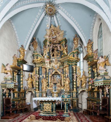 Zdjęcie nr 2: Nastawa ołtarzowa architektoniczna, trójosiowa z bramkami, dwukondygnacyjna, na dwustrefowym cokole. Ołtarz kamienny, prostopadłościenny, w drewnianej obudowie. Antepedium w formie wydłużonego, leżącego prostokąta, w polu obraz Ostatniej Wieczerzy. Mensa drewniana. Osie nastawy wydzielone parą półfilarów ujętych dwiema parami kolumn o kapitelach kompozytowych i skręconych trzonach. Dźwigają one pełne belkowanie silnie wyłamane na osi filarów, z mocno wysuniętym gzymsem, na skraju belkowania przerwany półkolisty przyczółek zwrócony na zewnątrz, na nim cokoły z figurami św. Władysława i św. Stefana. Na tle filarów ustawione figury św. Augustyna i św. Wojciecha, na bramkach rzeźby św. Marcina i św. Jana Jałmużnika. W polu głównym głęboka nisza w formie kwadratu zamkniętego łukiem falistym nadwieszonym, wewnątrz rzeźbiarska scena „Zabójstwa św. Stanisława”. Druga kondygnacja ujęta półfilarami, na tle których znajdują się anioły-kariatydy siedzące na cokołach z wolutami, podtrzymujące imposty. W polu prześwit w formie stojącego prostokąta z wpisanym krzyżem w kształcie litery „X”, przed którym znajduje się figura św. Andrzeja. Całość zwieńczona półpostacią Boga Ojca wśród obłoków i uskrzydlonych główek anielskich w glorii promienistej. Struktura obficie dekorowana ornamentem roślinnym, w partii pierwszej i drugiej kondygnacji uszy utworzone z akantu i wstęgi. Ołtarz flankują dodatkowo dwie pary wolnostojących półfilarów z półkolumnami o skręconych trzonach przed nimi, stojących na dwustrefowych cokołach, dźwigających imposty z figurami. Na zewnętrznej, niższej parze znajdują się figury św. Jana Ewangelisty i św. Jana Chrzciciela, na wewnętrznej, wyższej parze – św. Piotra i św. Pawła. Struktura polichromowana w sposób naśladujący marmoryzację w kolorze niebiesko-szarym, z zielonymi profilami oraz złoconymi kolumnami, profilami i ornamentyką.


