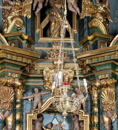 Zdjęcie nr 6: Nastawa ołtarzowa architektoniczna, trójosiowa z bramkami, dwukondygnacyjna, na dwustrefowym cokole. Ołtarz kamienny, prostopadłościenny, w drewnianej obudowie. Antepedium w formie wydłużonego, leżącego prostokąta, w polu obraz Ostatniej Wieczerzy. Mensa drewniana. Osie nastawy wydzielone parą półfilarów ujętych dwiema parami kolumn o kapitelach kompozytowych i skręconych trzonach. Dźwigają one pełne belkowanie silnie wyłamane na osi filarów, z mocno wysuniętym gzymsem, na skraju belkowania przerwany półkolisty przyczółek zwrócony na zewnątrz, na nim cokoły z figurami św. Władysława i św. Stefana. Na tle filarów ustawione figury św. Augustyna i św. Wojciecha, na bramkach rzeźby św. Marcina i św. Jana Jałmużnika. W polu głównym głęboka nisza w formie kwadratu zamkniętego łukiem falistym nadwieszonym, wewnątrz rzeźbiarska scena „Zabójstwa św. Stanisława”. Druga kondygnacja ujęta półfilarami, na tle których znajdują się anioły-kariatydy siedzące na cokołach z wolutami, podtrzymujące imposty. W polu prześwit w formie stojącego prostokąta z wpisanym krzyżem w kształcie litery „X”, przed którym znajduje się figura św. Andrzeja. Całość zwieńczona półpostacią Boga Ojca wśród obłoków i uskrzydlonych główek anielskich w glorii promienistej. Struktura obficie dekorowana ornamentem roślinnym, w partii pierwszej i drugiej kondygnacji uszy utworzone z akantu i wstęgi. Ołtarz flankują dodatkowo dwie pary wolnostojących półfilarów z półkolumnami o skręconych trzonach przed nimi, stojących na dwustrefowych cokołach, dźwigających imposty z figurami. Na zewnętrznej, niższej parze znajdują się figury św. Jana Ewangelisty i św. Jana Chrzciciela, na wewnętrznej, wyższej parze – św. Piotra i św. Pawła. Struktura polichromowana w sposób naśladujący marmoryzację w kolorze niebiesko-szarym, z zielonymi profilami oraz złoconymi kolumnami, profilami i ornamentyką.


