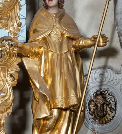 Zdjęcie nr 1: Figura ścięta z tyłu, ustawiona jest na obłoku, ukazana frontalnie z głową skierowaną w prawo, w kontrapoście, o esowato wygiętej sylwetce, z rękami rozłożonymi na boki, w lewej trzyma pastorał. Twarz ma owalną, pełną, o oczach wzniesionych w górę, ustach lekko rozchylonych, okoloną falowanymi, sięgającymi ramion włosami. Święty ubrany jest w strój biskupi: albę, rokietę i mucet, na szyi ma zawieszony pektorał, na dłoniach rękawiczki, a na głowie infułę. Ramiona okrywa kapa spięta na piersi, z rozwianą na prawy bok fałdą. Po lewej stronie świętego niewielka, klęcząca postać kalekiego mężczyzny, wspierającego się lewą ręką na kulach, z wyciągniętą ku górze prawą ręką. Polichromia naturalistyczna w partiach ciała, szaty i atrybuty pozłacane.