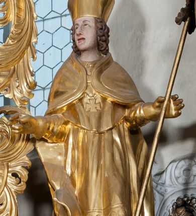 Zdjęcie nr 4: Figura ścięta z tyłu, ustawiona jest na obłoku, ukazana frontalnie z głową skierowaną w prawo, w kontrapoście, o esowato wygiętej sylwetce, z rękami rozłożonymi na boki, w lewej trzyma pastorał. Twarz ma owalną, pełną, o oczach wzniesionych w górę, ustach lekko rozchylonych, okoloną falowanymi, sięgającymi ramion włosami. Święty ubrany jest w strój biskupi: albę, rokietę i mucet, na szyi ma zawieszony pektorał, na dłoniach rękawiczki, a na głowie infułę. Ramiona okrywa kapa spięta na piersi, z rozwianą na prawy bok fałdą. Po lewej stronie świętego niewielka, klęcząca postać kalekiego mężczyzny, wspierającego się lewą ręką na kulach, z wyciągniętą ku górze prawą ręką. Polichromia naturalistyczna w partiach ciała, szaty i atrybuty pozłacane.