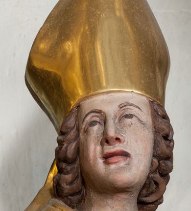Zdjęcie nr 5: Figura ścięta z tyłu, ustawiona jest na obłoku, ukazana frontalnie z głową skierowaną w prawo, w kontrapoście, o esowato wygiętej sylwetce, z rękami rozłożonymi na boki, w lewej trzyma pastorał. Twarz ma owalną, pełną, o oczach wzniesionych w górę, ustach lekko rozchylonych, okoloną falowanymi, sięgającymi ramion włosami. Święty ubrany jest w strój biskupi: albę, rokietę i mucet, na szyi ma zawieszony pektorał, na dłoniach rękawiczki, a na głowie infułę. Ramiona okrywa kapa spięta na piersi, z rozwianą na prawy bok fałdą. Po lewej stronie świętego niewielka, klęcząca postać kalekiego mężczyzny, wspierającego się lewą ręką na kulach, z wyciągniętą ku górze prawą ręką. Polichromia naturalistyczna w partiach ciała, szaty i atrybuty pozłacane.