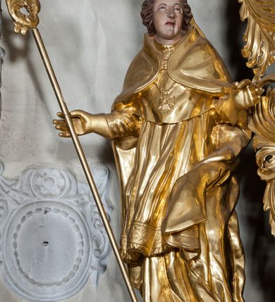 Zdjęcie nr 2: Figura ścięta z tyłu, stojąca na obłoku, ukazana frontalnie z głową zwróconą w lewą stronę, sylwetka esowato wygięta, ręce rozchylone na boki, w prawej trzyma pastorał. Twarz świętego owalna, pełna, oczy skierowane ku górze, usta lekko rozchylone, okolona krótkimi, falowanymi włosami, sięgającymi ramion. Marcin jest ubrany w szaty biskupie: albę, rokietę i mucet, na głowie ma infułę, na piersi zawieszony pektorał, na dłoniach rękawiczki. Ramiona okrywa kapa spięta na piersi, z rozwianą fałdą przy jego lewym boku. Obok świętego, po jego prawej, półpostać zwrócona w trzech czwartych w prawo, z rękoma złożonymi w geście modlitwy. Szaty postaci złocone, karnacja ciała w odcieniu jasnokremowym. 