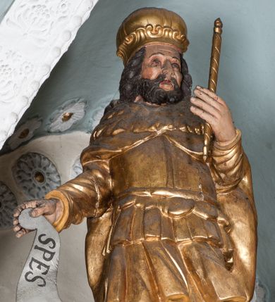 Zdjęcie nr 1: Figura jest drążona z tyłu, ustawiona na rzeźbionym obłoku, zwrócona frontalnie. Święty w lewej ręce wysuniętej przed siebie trzyma berło, w prawej wyciągniętej w bok banderolę z napisem „SPES MEA IN DEO”. Twarz ma kwadratową, o dużych oczach, niewielkim nosie i pełnych ustach, okoloną krótką, gęstą brodą oraz kręconymi włosami sięgającymi ramion. Ubrany jest w tunikę sięgającą ramion, zbroję torsową, spodnie i wysokie buty, na ramionach ma płaszcz z kołnierzem, na głowie koronę zamkniętą. Polichromia naturalistyczna w odsłoniętych partiach ciała, strój i atrybuty pozłocone.
