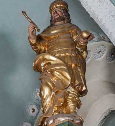 Zdjęcie nr 2: Figura ustawiona jest na rzeźbionym obłoku, zwrócona frontalnie, w kontrapoście, w lewej, wyciągniętej w bok ręce trzyma kulę (jabłko królewskie?), w prawej, wysuniętej w przód – berło. Twarz świętego kwadratowa, z oczami skierowanymi w górę, małym nosem i rozchylonymi ustami, okolona krótką brodą oraz sięgającymi ramion, kręconymi włosami. Stefan jest ubrany w suknię sięgającą kolan, zbroję torsową oraz płaszcz zawinięty na prawej nodze, spodnie i wysokie buty, na głowie ma koronę zamkniętą. Polichromia naturalistyczna w odsłoniętych partiach ciała, strój i atrybuty pozłacane.