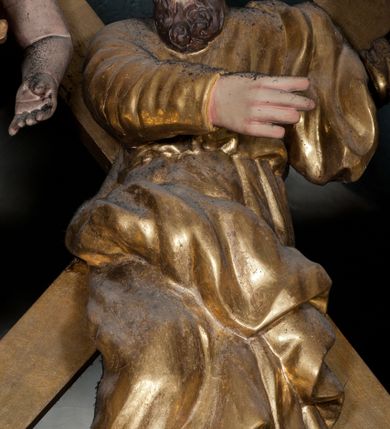 Zdjęcie nr 1: Rzeźba drążona, ustawiona na tle krzyża w formie litery „X” ujętego w profilowaną ramę. Figura zwrócona jest frontalnie, w kontrapoście, lewą ręką ujmuje ramię krzyża, prawą wyciąga przed siebie. Twarz ma pociągłą, nos prosty, oczy skierowane w dół, usta rozchylone, okoloną brodą skręconą w pukle oraz krótkimi włosami. Postać jest ubrana w suknię ściśle przylegającą do ciała, przepasaną w talii oraz płaszcz zarzucony na lewe ramię oraz opadający na kolana w postaci skręconej tkaniny. Polichromia naturalistyczna w odsłoniętych partiach ciała, strój i krzyż pozłocone. 
