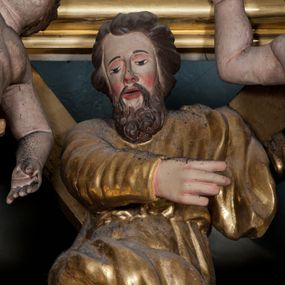 Zdjęcie nr 1: Rzeźba drążona, ustawiona na tle krzyża w formie litery „X” ujętego w profilowaną ramę. Figura zwrócona jest frontalnie, w kontrapoście, lewą ręką ujmuje ramię krzyża, prawą wyciąga przed siebie. Twarz ma pociągłą, nos prosty, oczy skierowane w dół, usta rozchylone, okoloną brodą skręconą w pukle oraz krótkimi włosami. Postać jest ubrana w suknię ściśle przylegającą do ciała, przepasaną w talii oraz płaszcz zarzucony na lewe ramię oraz opadający na kolana w postaci skręconej tkaniny. Polichromia naturalistyczna w odsłoniętych partiach ciała, strój i krzyż pozłocone. 