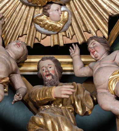 Zdjęcie nr 2: Rzeźba drążona, ustawiona na tle krzyża w formie litery „X” ujętego w profilowaną ramę. Figura zwrócona jest frontalnie, w kontrapoście, lewą ręką ujmuje ramię krzyża, prawą wyciąga przed siebie. Twarz ma pociągłą, nos prosty, oczy skierowane w dół, usta rozchylone, okoloną brodą skręconą w pukle oraz krótkimi włosami. Postać jest ubrana w suknię ściśle przylegającą do ciała, przepasaną w talii oraz płaszcz zarzucony na lewe ramię oraz opadający na kolana w postaci skręconej tkaniny. Polichromia naturalistyczna w odsłoniętych partiach ciała, strój i krzyż pozłocone. 