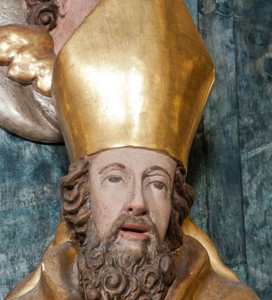 Zdjęcie nr 5: Rzeźba ścięta z tyłu, ustawiona jest na obłoku, zwrócona frontalnie, o esowato wygiętej sylwetce, z rękoma rozłożonymi na boki. Święty w lewej dłoni trzyma gorejące serce, w prawej pastorał. Twarz ma pociągłą o prostym nosie, głęboko rzeźbionej górnej powiece oraz szerokich, rozchylonych ustach, okoloną gęstą brodą, silnie kręconą oraz krótkimi włosami. Ubrany jest w albę, komżę, mucet i kapę, na piersi ma krzyż pektoralny, na głowie infułę, na dłoniach zaś rękawiczki. Polichromia naturalistyczna w odsłoniętych partiach ciała, strój i pastorał pozłocone, serce posrebrzone.