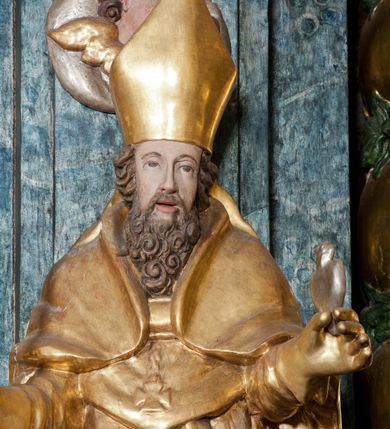 Zdjęcie nr 4: Rzeźba ścięta z tyłu, ustawiona jest na obłoku, zwrócona frontalnie, o esowato wygiętej sylwetce, z rękoma rozłożonymi na boki. Święty w lewej dłoni trzyma gorejące serce, w prawej pastorał. Twarz ma pociągłą o prostym nosie, głęboko rzeźbionej górnej powiece oraz szerokich, rozchylonych ustach, okoloną gęstą brodą, silnie kręconą oraz krótkimi włosami. Ubrany jest w albę, komżę, mucet i kapę, na piersi ma krzyż pektoralny, na głowie infułę, na dłoniach zaś rękawiczki. Polichromia naturalistyczna w odsłoniętych partiach ciała, strój i pastorał pozłocone, serce posrebrzone.