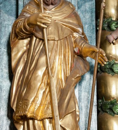 Zdjęcie nr 1: Rzeźba ścięta z tyłu, zwrócona jest w trzech czwartych w prawo, w kontrapoście, o esowato wygiętej sylwetce. Święty w lewej wyciągniętej w bok ręce trzyma pastorał, w prawej wiosło. Twarz ma pociągłą o prostym nosie, głęboko rzeźbionej górnej powiece, rozchylonych, pełnych ustach, okoloną brodą skręconą w dwa pukle oraz krótkimi włosami. Ubrany jest w albę, rokietę, mucet, kapę oraz paliusz, na głowie ma infułę, na dłoniach rękawiczki. Rzeźba polichromowana naturalistycznie w odsłoniętych partiach ciała, strój i atrybuty pozłocone.