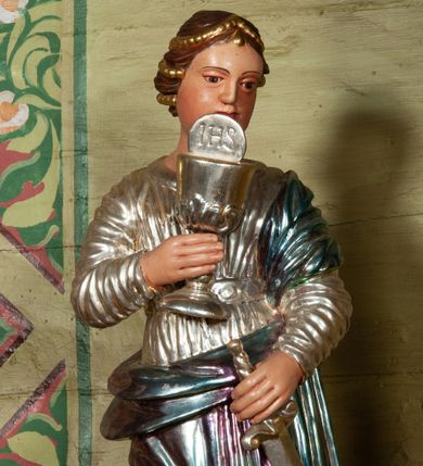 Zdjęcie nr 1: Rzeźba ścięta z tyłu, ustawiona na podstawie w formie rzeźbionego obłoku. Figura zwrócona frontalnie, w kontrapoście, z głową skierowaną w lewo. W prawej, ugiętej ręce trzyma przed sobą kielich z Hostią; w lewej miecz zwrócony ostrzem w dół, na zewnątrz. Twarz pociągła, pełna, o wydatnym podbródku, rysy delikatnie rzeźbione. Włosy ma upięte z tyłu głowy, z wplecionymi sznurami pereł. Ubrana jest w srebrzoną suknię z długim rękawem, przepasaną w talii, obficie drapowaną w drobne fałdki; oraz laserunkowy płaszcz przerzucony przez lewe ramie, otaczający postać i przewiązany na lewym biodrze, drapowany w równoległe fałdy z zawiniętym górnym brzegiem.Polichromia naturalistyczna w odsłoniętych partiach ciała. 