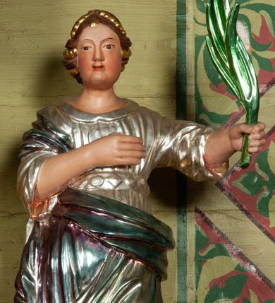Zdjęcie nr 1: Rzeźba ścięta z tyłu, drążona, ustawiona na podstawie w formie rzeźbionego obłoku. Figura zwrócona frontalnie. W lewej ręce wyciągniętej w bok trzyma palmę męczeństwa, prawa spoczywa na piersi. Twarz pociągła, o pełnych policzkach, wydatnym podbródku i delikatnie rzeźbionych rysach. Włosy upięte z tyłu głowy z wplecionymi sznurami pereł. Ubrana jest w srebrzoną suknię o szerokich rękawach, z kołnierzykiem, przepasaną w talii kameryzowanym pasem, gęsto drapowaną w równoległe, rurkowate fałdy; oraz laserunkowy, błękitny płaszcz, przewiązany przez biodra na prawym boku. U nóg świętej fragment koła. Polichromia naturalistyczna w odsłoniętych partiach ciała. 