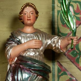 Zdjęcie nr 1: Rzeźba ścięta z tyłu, drążona, ustawiona na podstawie w formie rzeźbionego obłoku. Figura zwrócona frontalnie. W lewej ręce wyciągniętej w bok trzyma palmę męczeństwa, prawa spoczywa na piersi. Twarz pociągła, o pełnych policzkach, wydatnym podbródku i delikatnie rzeźbionych rysach. Włosy upięte z tyłu głowy z wplecionymi sznurami pereł. Ubrana jest w srebrzoną suknię o szerokich rękawach, z kołnierzykiem, przepasaną w talii kameryzowanym pasem, gęsto drapowaną w równoległe, rurkowate fałdy; oraz laserunkowy, błękitny płaszcz, przewiązany przez biodra na prawym boku. U nóg świętej fragment koła. Polichromia naturalistyczna w odsłoniętych partiach ciała. 