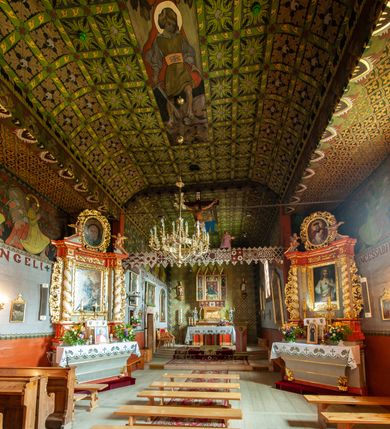 Zdjęcie nr 10: Polichromia pokrywa praktycznie całe wnętrze kościoła wraz z przedsionkiem. Przy wejściu, na stropie pod wieżą znajduje się polichromia patronowa. Jest wielobrawna, z motywami architektonicznymi, geometrycznymi, roślinno-kwiatowymi i figuralnymi – jeleniami, parami gołąbków, orłami, smokami. Wśród nich postać jeźdźca z kopią i grupa ludzi udających się na polowanie, poniżej których napis „Odbudowę kościoła, urządzenie wnętrza i rekonstrukcję dokonano staraniem Ks(iędza) Andrzeja Braszki / w czasie od lipca 1932 do grudnia r(oku) (19)39. / Pracę rozpoczął Władysław Cichoń + Skończył Adam Stalony-Dobrzański z Eugeniuszem Pisarkiem”. W polichromii przedsionka przeważają kolory  biały, czerwony, zielony, niebieski, żółty i czarny. Ponad wejściem do nawy napis  „ZACZNIJCIE / WARGI NASZE / CHWALIĆ PANNĘ / ŚWIĘTĄ / ZACZNIJCIE OPOWIADAĆ / CZEŚĆ JEJ NIEPOJĘTĄ”  i po drugieh stronie „PRZYBĄDŹ NAM. MIŁOŚCIWA PANI / KU POMOCY / A WYBAW NAS / Z RĄK POTĘŻNYCH / NIEPRZYJACIÓŁ MOCY”. Poniżej „CHWAŁA OJCU I SYNOWI JEGO PRZEDWIECZNEMU”.
W prezbiterium na ścianie i na suficie imitacja kasetonów z rozetami z przewagą koloru zielonego uzupełnionego czerwonym na ścianie i czarnym oraz bladożółtym na stropie. W centrum sufitu postać Matki Boskiej z Dzieciątkiem na lewym ramieniu, za którymi dwaj aniołowie skłaniający głowy. Maria ma poważną twarz o delikatnych rysach, ciemne oczy i wąski nos, na głowie otwartą koronę, wokół niej nimb. Jest ubrana w jasną sukienkę i intensywnie niebieski płaszcz. Dzieciątko prawą ręką błogosławi, lewą trzyma rękę Marii. Ma owalną, delikatną twarz, a na sobie jasną tunikę i zamkniętą koronę. 
W nawie, pośród takich samych kasetonów jak w prezbiterium, na sklepieniu wyobrażenie św. Michała Archanioła zabijającego szatana. Ponad nim postać anioła splatającego dłonie pod brodą. Archanioł spogląda pod stopy, w prawej ręce trzyma skierowany ostrzem w stronę potwora miecz, lewą ma opuszczoną do boku. Ma na sobie zieloną tunikę przewiązaną szerokim, góralskim pasem i czerwony płaszcz. Jasne postrzępione plamy ponad jego plecami sugerują skrzydła. Anioł powyżej jest ubrany w czerwoną sukienkę, nad nim płynnie uchodzą w górę ulistnione linie. Na ścianach nawy, pod linią stropu, rząd klęczących aniołów między kwitnącymi kwiatami. Każdy jest ubrany w zielonożółtą sukienkę i sandały, ma włosy do ramion i złoty nimb. Skrzydła z różnie ułożonych piór są wielobarwne. Anioły klęczą na linii ornamentu, ściana poniżej jest jasnoniebieska, z napisem powyżej opisanych przedstawień. 
Na skośnych fragmentach stropu imitacja drobniejszych kasetonów o falistym zarysie, wypełnionych krzyżami równoramiennymi. Wszystkie ciemnożółte, większość na czarnym tle, niektóre na tle zielonym tworzą kształt rombów. Na krawędzi motywy geometryczne, które mogą sugerować płonące serca. 
Na parapecie chóru fragmenty dawnej polichromii o motywach liściasto-wiciowych z rozwiniętymi kwiatami i motywem żołędzia. Między nimi ludzka głowa. Malowidło utrzymane w dominującym kolorze brązu, z elementami zielonymi i czerwonymi. 
