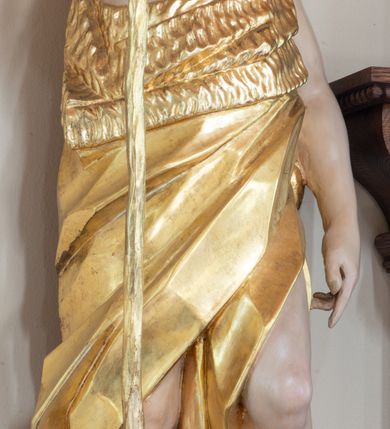 Zdjęcie nr 4: Święty ukazany frontalnie, z głową skierowaną w lewo, w prawej ręce trzyma laskę zakończoną krzyżem, lewą ma opuszczoną wzdłuż boku, lewą nogę opiera na rzeźbionym kamieniu, na którym stoi baranek. Twarz ma pociągłą, o migdałowatych oczach skierowanych w górę, prostym nosie i rozchylonych ustach, okoloną krótką, kręconą brodą oraz kręconymi włosami opadającymi na ramiona. Ubrany jest w melotę opasującą jego korpus i nogi, odsłaniającą część klatki piersiowej i ramiona oraz lewą nogę. Polichromia ciała naturalistyczna, włosy i broda brązowe, strój i laska złocone, baranek srebrzony. 