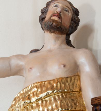 Zdjęcie nr 5: Święty ukazany frontalnie, z głową skierowaną w lewo, w prawej ręce trzyma laskę zakończoną krzyżem, lewą ma opuszczoną wzdłuż boku, lewą nogę opiera na rzeźbionym kamieniu, na którym stoi baranek. Twarz ma pociągłą, o migdałowatych oczach skierowanych w górę, prostym nosie i rozchylonych ustach, okoloną krótką, kręconą brodą oraz kręconymi włosami opadającymi na ramiona. Ubrany jest w melotę opasującą jego korpus i nogi, odsłaniającą część klatki piersiowej i ramiona oraz lewą nogę. Polichromia ciała naturalistyczna, włosy i broda brązowe, strój i laska złocone, baranek srebrzony. 