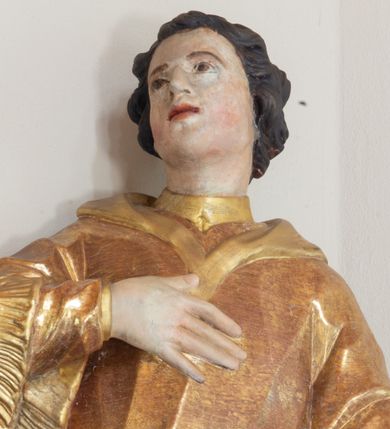 Zdjęcie nr 1: Święty zwrócony w trzech czwartych w prawo, lekko odchylony, prawą rękę kładzie na piersi, lewą wyciąga w bok,  lewa noga oparta na stosie kamieni. Twarz o młodzieńczych rysach, owalna, o migdałowatych oczach skierowanych w górę, prostym nosie i pełnych, rozchylonych ustach, okolona krótkimi i kręconymi włosami. Ubrany jest w albę oraz dalmatykę z frędzlą przy brzegach. Polichromia ciała naturalistyczna, włosy brązowe, strój złocony. 