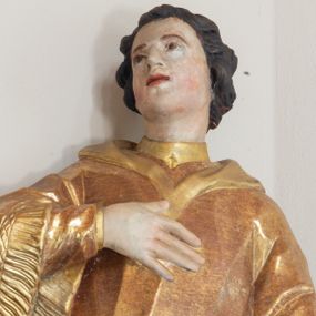 Zdjęcie nr 1: Święty zwrócony w trzech czwartych w prawo, lekko odchylony, prawą rękę kładzie na piersi, lewą wyciąga w bok,  lewa noga oparta na stosie kamieni. Twarz o młodzieńczych rysach, owalna, o migdałowatych oczach skierowanych w górę, prostym nosie i pełnych, rozchylonych ustach, okolona krótkimi i kręconymi włosami. Ubrany jest w albę oraz dalmatykę z frędzlą przy brzegach. Polichromia ciała naturalistyczna, włosy brązowe, strój złocony. 