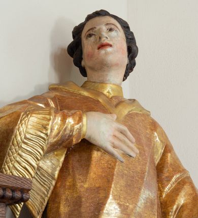 Zdjęcie nr 5: Święty zwrócony w trzech czwartych w prawo, lekko odchylony, prawą rękę kładzie na piersi, lewą wyciąga w bok,  lewa noga oparta na stosie kamieni. Twarz o młodzieńczych rysach, owalna, o migdałowatych oczach skierowanych w górę, prostym nosie i pełnych, rozchylonych ustach, okolona krótkimi i kręconymi włosami. Ubrany jest w albę oraz dalmatykę z frędzlą przy brzegach. Polichromia ciała naturalistyczna, włosy brązowe, strój złocony. 