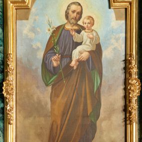 Zdjęcie nr 1: Podobrazie w formie stojącego prostokąta, pole obrazowe zamknięta łukiem półkolistym nadwieszonym. W centrum św. Józef ukazany w całej postaci, frontalnie, w wykroku, z głową przechyloną na lewe ramię. W prawej, ugiętej ręce trzyma gałązkę lilii, lewą podtrzymuje Dzieciątko. Twarz ma pociągłą, o delikatnych rysach, dużych oczach, prostym nosie i pełnych ustach, okoloną krótkimi, siwymi włosami i brodą. Ubrany jest w obszerną, fioletową tunikę, przepasaną w talii, ze złoconą lamówką wokół otworu na szyję. Na ramiona ma narzucony brązowy płaszcz z zieloną podszewką, który podtrzymuje z przodu lewą dłonią. Na stopach ma sandały. Dzieciątko ukazane w pozycji siedzącej, zwrócone w trzech czwartych w lewo, z głową przedstawioną frontalnie. Twarz ma okrągłą, o dużych oczach, małym nosie i pełnych ustach, okoloną krótkimi, jasnymi włosami. Ubrane jest w białą, luźną tunikę ze złoconą lamówką wokół otworu na szyję. Postacie ukazane na tle nieba wypełnionego błekitno-szarymi obłokami.


