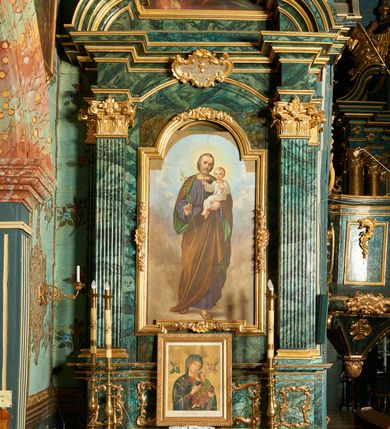 Zdjęcie nr 2: Podobrazie w formie stojącego prostokąta, pole obrazowe zamknięta łukiem półkolistym nadwieszonym. W centrum św. Józef ukazany w całej postaci, frontalnie, w wykroku, z głową przechyloną na lewe ramię. W prawej, ugiętej ręce trzyma gałązkę lilii, lewą podtrzymuje Dzieciątko. Twarz ma pociągłą, o delikatnych rysach, dużych oczach, prostym nosie i pełnych ustach, okoloną krótkimi, siwymi włosami i brodą. Ubrany jest w obszerną, fioletową tunikę, przepasaną w talii, ze złoconą lamówką wokół otworu na szyję. Na ramiona ma narzucony brązowy płaszcz z zieloną podszewką, który podtrzymuje z przodu lewą dłonią. Na stopach ma sandały. Dzieciątko ukazane w pozycji siedzącej, zwrócone w trzech czwartych w lewo, z głową przedstawioną frontalnie. Twarz ma okrągłą, o dużych oczach, małym nosie i pełnych ustach, okoloną krótkimi, jasnymi włosami. Ubrane jest w białą, luźną tunikę ze złoconą lamówką wokół otworu na szyję. Postacie ukazane na tle nieba wypełnionego błekitno-szarymi obłokami.


