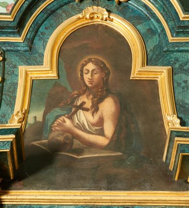 Zdjęcie nr 1: Obraz w formie trapezu zamkniętego łukiem półkolistym nadwieszonym, o wklęsłych bokach. W centrum ukazana półpostać św. Marii Magdaleny w jaskini. Święta zwrócona w trzech czwartych w prawo, ze splecionymi dłońmi trzymającymi krzyż, opartymi na czaszce leżącej na otwartej księdze. Twarz ma owalną, o dużych, okrągłych oczach skierowany w dół, wąskim, długim nosie i pełnych ustach. Włosy ma długie, falowane, opadające na ramiona i plecy. Ubrana jest w białą szatę przewiązaną diagonalnie przez prawe ramię, odsłaniającą pierś oraz niebieski płaszcz zarzucony na plecy i prawe ramię. Święta ukazana na tle groty, z widocznym po lewej stronie górzystym pejzażem z wieżą. 




