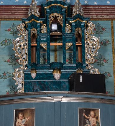 Zdjęcie nr 2: Szafa architektoniczna, pięcioosiowa, na profilowanym cokole, podtrzymywanym przez trzy wsporniki zakończone szyszkami. Oś środkowa najwyższa, dwie skrajne niższe, ujęte pilastrami podtrzymującymi fragmenty belkowania wyłamanego półkoliście i pod kątem, z prześwitami na piszczałki w formie stojącego prostokąta. Dwie środkowe osie najniższe, zakończone fragmentami niepełnego belkowania, z prześwitami na piszczałki w formie stojącego prostokąta. Oś środkowa i dwie skrajne zwieńczone ornamentalnymi małżowinowo-chrząstkowymi szczytami. Na belkowaniu osi środkowych fragmenty ornamentu małżowinowo-chrząstkowego, którym zdobi również prześwity na piszczałki w górnej części. Szafa i jej cokół flankowane rozłożystymi uszami utworzonymi z ornamentu małżowinowo-chrząstkowego z motywami owoców i winnego grona. Struktura polichromowana na sposób imitujący marmoryzację w kolorze niebieskim, ornamentyka złocona i srebrzona.
