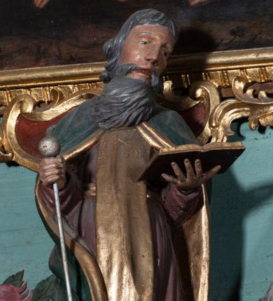 Zdjęcie nr 1: Figura ścięta z tyłu, ustawiona na niskiej podstawie. Święty ukazany frontalnie, z głową przechyloną na lewe ramię, o delikatnie esowato wygiętej sylwetce. Obie ręce ma wyciągnięte przed siebie, lewą podtrzymuje otwartą księgę, w prawej trzyma laskę zakończoną kulą. Twarz ma pociągłą, o wąskich oczach i nosie oraz rozchylonych ustach, okoloną długą, szarą brodą rozwianą w bok oraz krótkimi, siwymi włosami. Ubrany jest w brązową tunikę przepasaną w talii, ze złoconym szkaplerzem oraz zielony płaszcz ze złoconą podszewką, zarzucony na ramiona i zapięty pod szyją. Polichromia ciała naturalistyczna.

