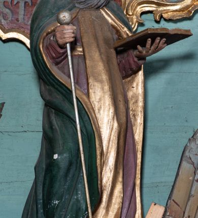 Zdjęcie nr 2: Figura ścięta z tyłu, ustawiona na niskiej podstawie. Święty ukazany frontalnie, z głową przechyloną na lewe ramię, o delikatnie esowato wygiętej sylwetce. Obie ręce ma wyciągnięte przed siebie, lewą podtrzymuje otwartą księgę, w prawej trzyma laskę zakończoną kulą. Twarz ma pociągłą, o wąskich oczach i nosie oraz rozchylonych ustach, okoloną długą, szarą brodą rozwianą w bok oraz krótkimi, siwymi włosami. Ubrany jest w brązową tunikę przepasaną w talii, ze złoconym szkaplerzem oraz zielony płaszcz ze złoconą podszewką, zarzucony na ramiona i zapięty pod szyją. Polichromia ciała naturalistyczna.

