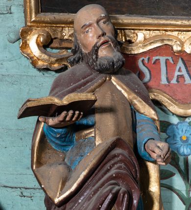 Zdjęcie nr 1: Figura ścięta z tyłu, ustawiona na niskiej podstawie. Święty ukazany frontalnie, z głową przechyloną na prawe ramię, obie ręce wyciąga do przodu, prawą podtrzymuje otwartą księgę. Twarz ma pociągłą, o wąskich oczach i nosie oraz otwartych ustach, okoloną gęstą brodą oraz włosami sięgającymi ramion, na głowie ma łysinę. Ubrany jest w niebieską suknię, przepasaną w talii, ze złoconym szkaplerzem oraz brązowy płaszcz o złoconej podszewce, zarzucony na ramiona, zapięty pod szyją i zawinięty z przodu postaci. Polichromia ciała naturalistyczna. 

