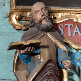 Zdjęcie nr 1: Figura ścięta z tyłu, ustawiona na niskiej podstawie. Święty ukazany frontalnie, z głową przechyloną na prawe ramię, obie ręce wyciąga do przodu, prawą podtrzymuje otwartą księgę. Twarz ma pociągłą, o wąskich oczach i nosie oraz otwartych ustach, okoloną gęstą brodą oraz włosami sięgającymi ramion, na głowie ma łysinę. Ubrany jest w niebieską suknię, przepasaną w talii, ze złoconym szkaplerzem oraz brązowy płaszcz o złoconej podszewce, zarzucony na ramiona, zapięty pod szyją i zawinięty z przodu postaci. Polichromia ciała naturalistyczna. 

