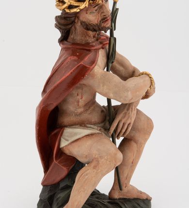 Zdjęcie nr 2: Rzeźba pełnoplastyczna, na podstawie w formie pnia. Chrystus ukazany w pozycji siedzącej, frontalnie, przechylony w prawo, z wysuniętą w bok prawą nogą. Ręce ma ugięte i lekko uniesione, skrzyżowane w nadgarstkach i przewiązane powrozem. Rękami obejmuje trzcinę. Twarz ma trójkątną, o migdałowatych oczach, dużym nosie i pełnych ustach; okoloną krótką brodą oraz silnie skręconymi po bokach głowy włosami, opadającymi na kark. Na głowie ma złoconą koronę cierniową. W partii bioder przewiązany jest białą tkaniną. Na ramiona ma narzucony czerwony płaszcz zawiązany pod szyją. Jego ciało jest szczupłe, ale muskularne. Polichromia ciała naturalistyczna z zaznaczonymi śladami męki. 

