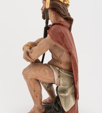 Zdjęcie nr 5: Rzeźba pełnoplastyczna, na podstawie w formie pnia. Chrystus ukazany w pozycji siedzącej, frontalnie, przechylony w prawo, z wysuniętą w bok prawą nogą. Ręce ma ugięte i lekko uniesione, skrzyżowane w nadgarstkach i przewiązane powrozem. Rękami obejmuje trzcinę. Twarz ma trójkątną, o migdałowatych oczach, dużym nosie i pełnych ustach; okoloną krótką brodą oraz silnie skręconymi po bokach głowy włosami, opadającymi na kark. Na głowie ma złoconą koronę cierniową. W partii bioder przewiązany jest białą tkaniną. Na ramiona ma narzucony czerwony płaszcz zawiązany pod szyją. Jego ciało jest szczupłe, ale muskularne. Polichromia ciała naturalistyczna z zaznaczonymi śladami męki. 

