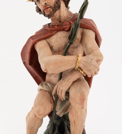 Zdjęcie nr 4: Rzeźba pełnoplastyczna, na podstawie w formie pnia. Chrystus ukazany w pozycji siedzącej, frontalnie, przechylony w prawo, z wysuniętą w bok prawą nogą. Ręce ma ugięte i lekko uniesione, skrzyżowane w nadgarstkach i przewiązane powrozem. Rękami obejmuje trzcinę. Twarz ma trójkątną, o migdałowatych oczach, dużym nosie i pełnych ustach; okoloną krótką brodą oraz silnie skręconymi po bokach głowy włosami, opadającymi na kark. Na głowie ma złoconą koronę cierniową. W partii bioder przewiązany jest białą tkaniną. Na ramiona ma narzucony czerwony płaszcz zawiązany pod szyją. Jego ciało jest szczupłe, ale muskularne. Polichromia ciała naturalistyczna z zaznaczonymi śladami męki. 

