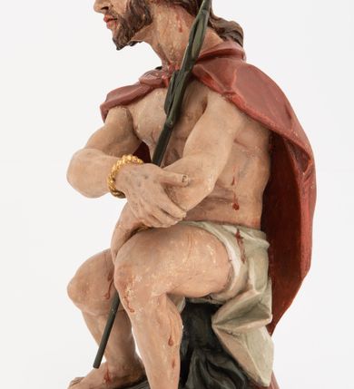 Zdjęcie nr 3: Rzeźba pełnoplastyczna, na podstawie w formie pnia. Chrystus ukazany w pozycji siedzącej, frontalnie, przechylony w prawo, z wysuniętą w bok prawą nogą. Ręce ma ugięte i lekko uniesione, skrzyżowane w nadgarstkach i przewiązane powrozem. Rękami obejmuje trzcinę. Twarz ma trójkątną, o migdałowatych oczach, dużym nosie i pełnych ustach; okoloną krótką brodą oraz silnie skręconymi po bokach głowy włosami, opadającymi na kark. Na głowie ma złoconą koronę cierniową. W partii bioder przewiązany jest białą tkaniną. Na ramiona ma narzucony czerwony płaszcz zawiązany pod szyją. Jego ciało jest szczupłe, ale muskularne. Polichromia ciała naturalistyczna z zaznaczonymi śladami męki. 

