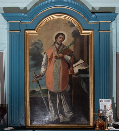 Zdjęcie nr 2: Obraz w formie stojącego prostokąta zamkniętego łukiem półkolistym nadwieszonym. W centrum św. Walenty ukazany w całej postaci, frontalnie, z głową skierowaną w lewo. W prawej opuszczonej ręce trzyma rękojeść miecza, skierowanego ostrzem w dół, lewą kładzie na piersi, w dłoni trzyma liść palmy. Twarz ma pociągłą, o małych, okrągłych oczach i uniesionych łukowatych brwiach, lekko garbatym nosie i małych, pełnych ustach; okoloną krótkimi, ciemnobrązowymi włosami. Ubrany jest w białą albę o brzegach dekorowanych koronką oraz jasnoróżowy ornat z jasnoniebieską kolumną o roślinnym wzorze. Wokół głowy ma nimb promienisty. Po lewej stronie świętego stół nakryty niebieską tkaniną, na którym leży otwarta księga i czarny biret, obok widoczna kolumna. Święty ukazany na tle górzystego pejzażu, w górnej części obrazu trzy uskrzydlone główki anielskie. 