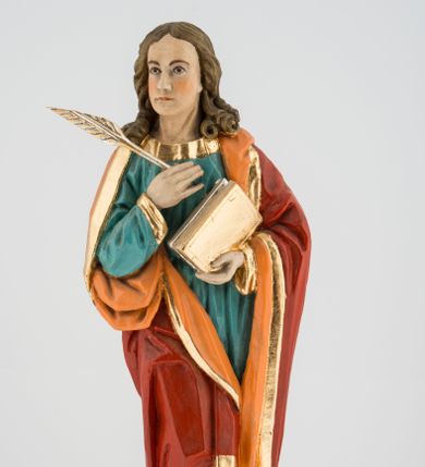Zdjęcie nr 1: Rzeźba pełna na cokole przedstawiająca św. Jana Ewangelistę. Święty ukazany jest w pozycji stojącej, frontalnie, w kontrapoście, z prawą nogą zgiętą w kolanie i głową skierowaną delikatnie w prawo. Prawą rękę przyciska do piersi, trzymając pióro, lewą przytrzymuje zamkniętą księgę. Ma pociągłą twarz o szeroko otwartych, brązowych oczach, długim nosie i wąskich ustach, okoloną długimi włosami zwijającymi się na końcach w pukle, z przedziałkiem na środku. Ubrany jest w zieloną suknię lamowaną przy krawędziach złotem oraz czerwony, podbity pomarańczowym materiałem płaszcz zarzucony na oba ramiona, układający się w głębokie fałdy. Stopy bose, przy lewej nodze świętego widoczny fragment niewielkiej postaci orła w biało-brązowe cętki, z pomarańczowym dziobem. Polichromia w partiach ciała naturalistyczna, księga i pióro złocone. 