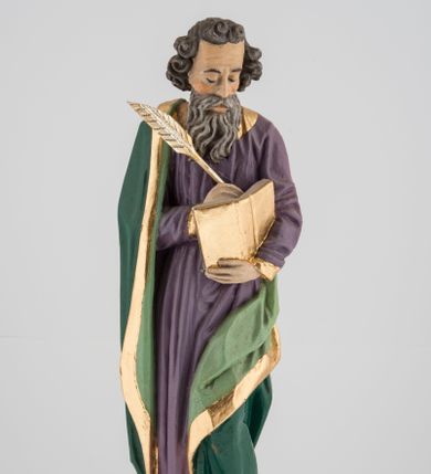 Zdjęcie nr 1: Rzeźba pełna na cokole, przedstawiająca św. Łukasza Ewangelistę. Święty ukazany został w pozycji stojącej, z górną partią ciała skręconą nieznacznie w lewo, w stronę trzymanej w lewej ręce księgi, w której pisze piórem trzymanym w prawej, obie ręce ugięte. Ma pociągłą twarz z przymkniętymi oczami, długim nosem i wąskimi ustami, okoloną wyraźnie zwiniętymi puklami włosów oraz długą, dzielącą się na pasma brodę i wąsy. Święty jest ubrany w przewiązaną w pasie fioletową suknię ze złoconymi lamówkami oraz ciemnozielony płaszcz podbity jasnozielonym materiałem, zarzucony na prawe ramię i otaczający lewe udo, układający się w głębokie fałdy. Stopy bose, przy prawej nodze niewielka, widoczna częściowo postać leżącego woła. Polichromia w partiach ciała naturalistyczna, byk ciemnobrązowy, księga, pióro i rogi byka złocone. 