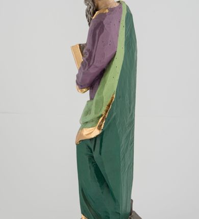 Zdjęcie nr 3: Rzeźba pełna na cokole, przedstawiająca św. Łukasza Ewangelistę. Święty ukazany został w pozycji stojącej, z górną partią ciała skręconą nieznacznie w lewo, w stronę trzymanej w lewej ręce księgi, w której pisze piórem trzymanym w prawej, obie ręce ugięte. Ma pociągłą twarz z przymkniętymi oczami, długim nosem i wąskimi ustami, okoloną wyraźnie zwiniętymi puklami włosów oraz długą, dzielącą się na pasma brodę i wąsy. Święty jest ubrany w przewiązaną w pasie fioletową suknię ze złoconymi lamówkami oraz ciemnozielony płaszcz podbity jasnozielonym materiałem, zarzucony na prawe ramię i otaczający lewe udo, układający się w głębokie fałdy. Stopy bose, przy prawej nodze niewielka, widoczna częściowo postać leżącego woła. Polichromia w partiach ciała naturalistyczna, byk ciemnobrązowy, księga, pióro i rogi byka złocone. 