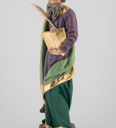 Zdjęcie nr 5: Rzeźba pełna na cokole, przedstawiająca św. Łukasza Ewangelistę. Święty ukazany został w pozycji stojącej, z górną partią ciała skręconą nieznacznie w lewo, w stronę trzymanej w lewej ręce księgi, w której pisze piórem trzymanym w prawej, obie ręce ugięte. Ma pociągłą twarz z przymkniętymi oczami, długim nosem i wąskimi ustami, okoloną wyraźnie zwiniętymi puklami włosów oraz długą, dzielącą się na pasma brodę i wąsy. Święty jest ubrany w przewiązaną w pasie fioletową suknię ze złoconymi lamówkami oraz ciemnozielony płaszcz podbity jasnozielonym materiałem, zarzucony na prawe ramię i otaczający lewe udo, układający się w głębokie fałdy. Stopy bose, przy prawej nodze niewielka, widoczna częściowo postać leżącego woła. Polichromia w partiach ciała naturalistyczna, byk ciemnobrązowy, księga, pióro i rogi byka złocone. 