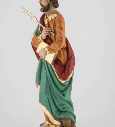 Zdjęcie nr 5: Rzeźba pełna, postać umieszczona na cokole przedstawiająca św. Marka Ewangelistę. Święty ukazany jest w pozycji stojącej, frontalnie, w kontrapoście, z prawą nogą zgiętą w kolanie i głową skierowaną na wprost. Prawą rękę przyciska do piersi, trzymając pióro, lewą przytrzymuje zamkniętą księgę. Święty ma pociągłą twarz o szeroko otwartych, niebieskich oczach, długim nosie i małych ustach, okoloną zwiniętymi w pukle włosami zakrywającymi uszy i formującymi lok nad czołem oraz wąsy i krótką brodę. Ubrany jest w jasnobrązową suknię lamowaną przy krawędziach złotem oraz w zielony, podbity czerwienią i lamowany złotem płaszcz przechodzący przez prawy bark i otaczający lewe biodro, układający się w głębokie fałdy. Stopy bose. Przy lewej nodze świętego widoczny fragment niewielkiej postaci lwa. Polichromia w partiach ciała naturalistyczna. 