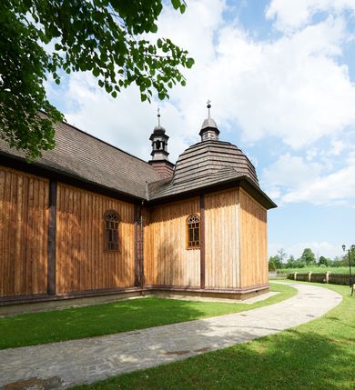 Zdjęcie nr 8: Kościół drewniany o konstrukcji zrębowej, na betonowym podmurowaniu przesłoniętym daszkiem gontowym. Elewacje jednolite, szalowane z listwowaniem, wzmocnione lisicami. Więźba dachowa drewniana, krokwiowa, o systemie więźbowo-zaskrzynieniowym. Kościół orientowany, jednonawowy, nawa na rzucie wydłużonego prostokąta, prezbiterium nieco węższe, zakończone trójbocznie. Do północnej ściany prezbiterium i nawy przylega zakrystia połączona z aneksem na rzucie prostokąta, do nawy od południa przylega duża kaplica na planie prostokąta otwarta do nawy otworem o wykroju prostokąta zamkniętego łukiem odcinkowym. 
Ściany wnętrza podzielone trzema parami pilastrów. Pomiędzy nawą a prezbiterium arkadowy otwór tęczowy z prostą belką. Parapet chóru muzycznego w zachodniej części nawy, oparty na dwóch kolumnach o wielobocznych trzonach. Wnętrze nakryte stropem płaskim, w nawie z zaskrzynieniami podtrzymywanymi przez trzy pary słupów o wielobocznych i kręconych trzonach. Otwory drzwiowe o prostokątnym wykroju zamknięte drzwiami jednoskrzydłowymi w północnej ścianie nawy oraz prezbiterium. Posadzka z płyt kamiennych. 
Elewacja frontowa jednoosiowa, zamknięta trójkątnym szczytem rozszerzającym się ku dołowi. Na osi ganek oparty na czterech słupach, nakryty dwuspadowym daszkiem gontowym, wejście do kościoła z dwuskrzydłowymi drzwiami. W partii szczytu dwa okna w formie stojącego prostokąta zamkniętego łukiem półkolistym. W elewacji południowej cztery otwory okienne w formie stojącego prostokąta zamkniętego łukiem półkolistym, dwa w nawie i dwa w prezbiterium oraz dwa analogiczne okna na zachodniej i wschodniej ścianie kaplicy. Elewacja północna z dwoma otworami okiennymi w formie stojącego prostokąta zamkniętego łukiem półkolistym, trzema oknami i wejściem do zakrystii i aneksu. Kościół nakryty wspólnym dachem o jednej kalenicy nad prezbiterium i nawą, gontowym, dwuspadowym, z przedłużoną połacią przekrywającą zaskrzynienia. W jednej trzeciej długości dachu nad nawą od strony prezbiterium wznosi się ponad kalenicą wieżyczka na sygnaturkę, na ośmiobocznym obitym blachą trzonie, z latarnią zwieńczoną niewielkim cebulastym hełmem. Kaplica boczna nakryta kopułą krytą gontem, z latarnią. Nad zakrystią i aneksem dach jednospadowy.
 
